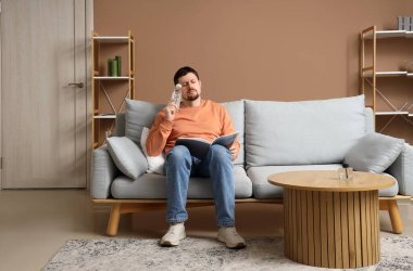 Evdeki kanepede su okuma dergisi okuyan yorgun genç bir adam.