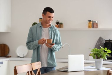 Masasında modern dizüstü bilgisayarı olan mutfakta kahve içen yakışıklı bir genç.