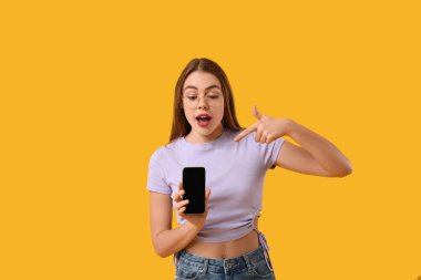 Şok olmuş genç bir kadın sarı arka planda cep telefonunu işaret ediyor.