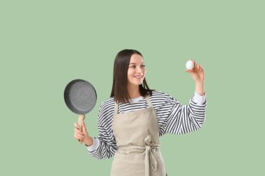 Yeşil arka planda tavuk yumurtası ve kızartma tavası olan önlüklü güzel bir kadın.