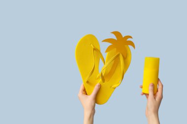 Mavi arka planda kağıt avuç içi ve güneş kremi spreyli sarı parmak arası terlikler tutan eller.