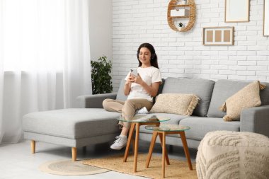 Oturma odasındaki gri koltukta cep telefonu kullanan kulaklıklı genç bir kadın.