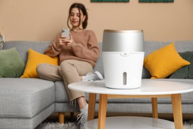 Moda oturma odasındaki modern nemlendiricinin yanındaki rahat koltukta cep telefonu kullanan genç bir kadın.