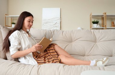 Güzel genç bir kadın kitap okuyor ve evdeki kanepede dinleniyor.