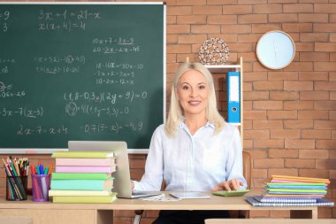 Sınıftaki masada dizüstü bilgisayar ve hesap makinesiyle çalışan kadın matematik öğretmeni