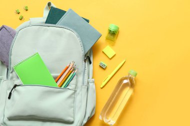 Okul çantasının içinde farklı kırtasiye malzemeleri ve sarı arka planda bir şişe su var.