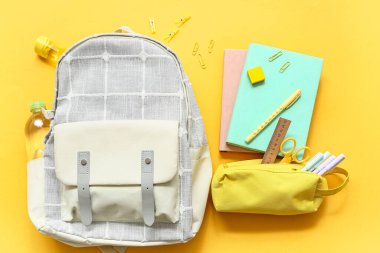 Farklı kırtasiye malzemeleriyle modern okul çantası ve sarı arka planda bir şişe su.