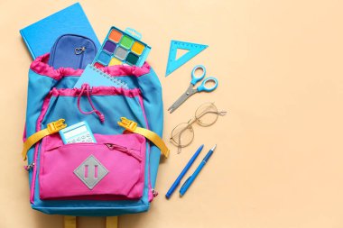 Bej arka planda farklı kırtasiye malzemeleri ve gözlüklerle modern okul sırt çantası.