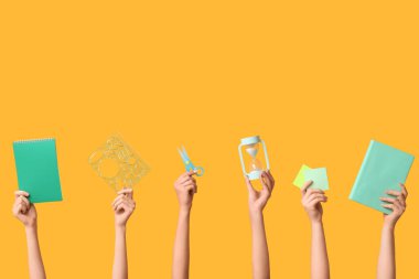 Sarı zemin üzerinde kırtasiye malzemesi, not defteri ve kum saati olan birçok el.