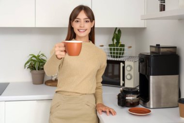 Güzel, genç, mutlu bir kadın, elinde bir fincan kahveyle mutfakta mola veriyor.