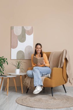 Kitap okuyan ve oturma odasının içindeki bej duvarın yanındaki koltukta oturan genç bir kadın.