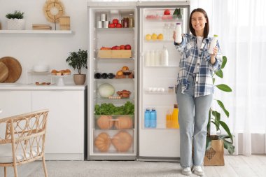 Mutfaktaki açık buzdolabının yanında yoğurt şişeleri olan genç bir kadın.