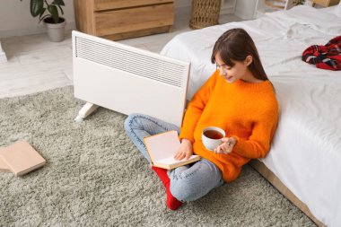 Yatak odasındaki elektrikli ısıtıcının yanında çay kitabı okuyan genç bir kadın.