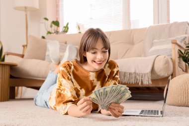 Evde yerde yatan dolar banknotları ve dizüstü bilgisayarı olan genç bir kadın.