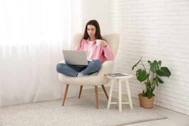 Evdeki ışık perdesinin yanındaki koltukta dizüstü bilgisayar kullanan genç bir kadın.