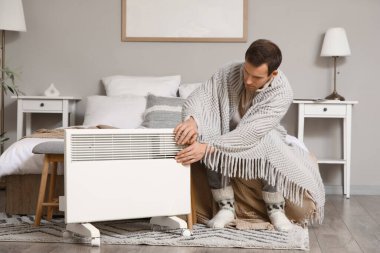 Yatak odasındaki elektrikli konteyner ısıtıcısının yanında ekoseli elleri olan genç bir adam.