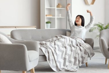 Evdeki kanepeye uzanan sıcak battaniyeli genç Asyalı kadın.