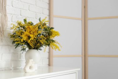 Beyaz tuğla duvarın yanındaki rafta mimoza çiçekleri olan vazo.