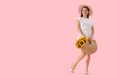Yazlık şapkalı şık genç kadın elinde hasır çantasıyla pembe arka planda ayçiçekleri tutuyor.