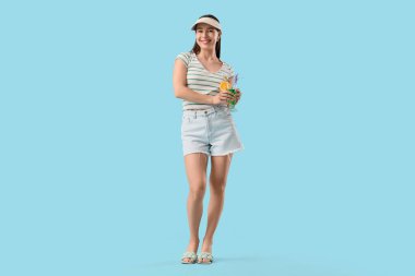 Mavi arka planda kokteyl eşliğinde yaz şapkalı şık bir genç kadın.