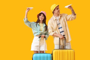 Sarı arka planda kağıt uçakları ve bavulları olan iki turist.