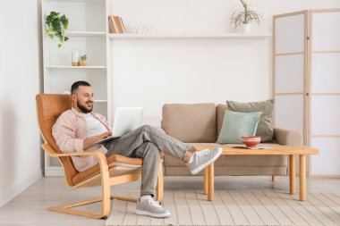 Evdeki kahverengi koltukta dizüstü bilgisayar kullanan yakışıklı adam.