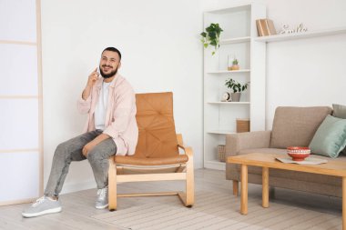 Evdeki kahverengi koltukta cep telefonuyla konuşan yakışıklı adam.