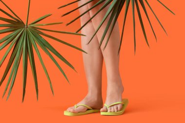 Turuncu arka planda palmiye yaprakları olan şık parmak arası terlikler giyen genç kadınların bacakları.