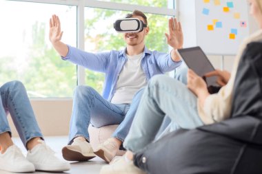 Genç adam, ofisteki puf minderlerinde iş arkadaşlarıyla VR gözlüğü kullanıyor.