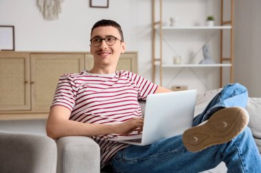 Gözlüklü genç adam evdeki koltukta dizüstü bilgisayar kullanıyor.