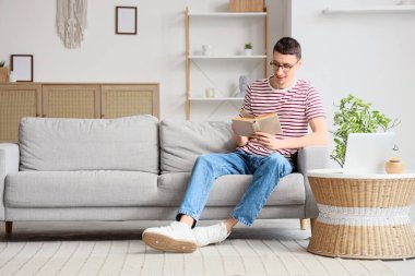 Gözlüklü genç adam evdeki kanepede kitap okuyor.