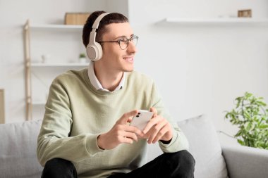 Gözlüklü, kulaklıklı, evdeki koltukta cep telefonu kullanan genç bir adam.