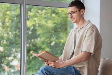 Gözlüklü genç adam evinin penceresinin yanında kitap okuyor.