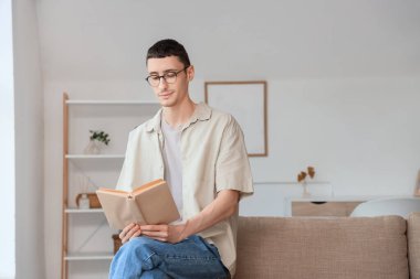 Gözlüklü genç adam evdeki kanepede kitap okuyor.