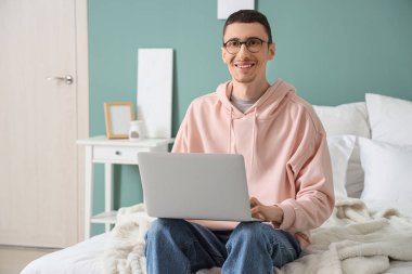 Yatak odasında dizüstü bilgisayar kullanan gözlüklü genç adam.