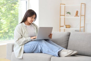 Evdeki kanepede dizüstü bilgisayar kullanan güzel genç bir kadın.