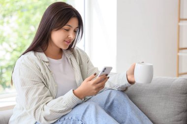 Evde cep telefonu kullanan, kahve fincanı olan güzel genç bir kadın.