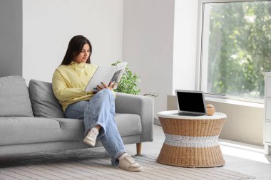 Güzel genç kadın evde kanepenin üzerinde dergi okuma