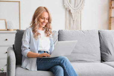 Evdeki kanepede dizüstü bilgisayar kullanan güzel sarışın kadın.