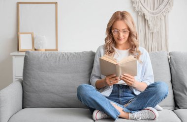 Evdeki kanepede kitap okuyan güzel sarışın kadın.