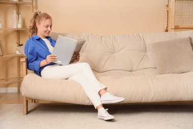 Güzel sarışın kadın evdeki kanepede dergi okuyor.