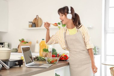 Kulaklıklı genç bir kadın mutfakta sebze salatası yiyor.