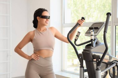 Spor salonunda spor bisikletinin yanında VR gözlüklü Afro-Amerikalı bir kadın.