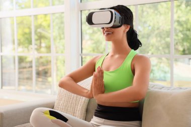 VR gözlüklü Afro-Amerikan kadın evdeki kanepede meditasyon yapıyor.
