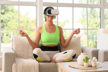 VR gözlüklü Afro-Amerikan kadın evdeki kanepede meditasyon yapıyor.