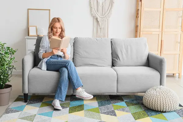 Evdeki kanepede kitap okuyan güzel sarışın kadın.