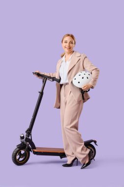 Leylak arka planında elektrikli scooter ve miğferi olan olgun bir iş kadını.