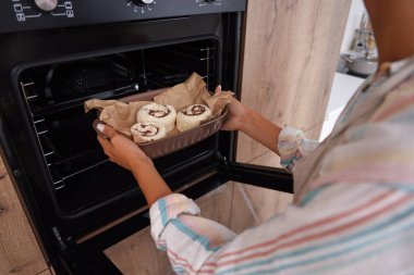 Genç bir kadın mutfaktaki fırına haşhaşlı çörek tepsisi koyuyor.