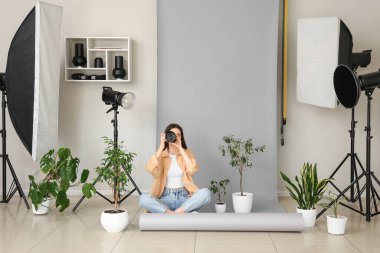 Fotoğraf stüdyosunda gri arka planda oturan kameralı ve çiçekli kadın fotoğrafçı.