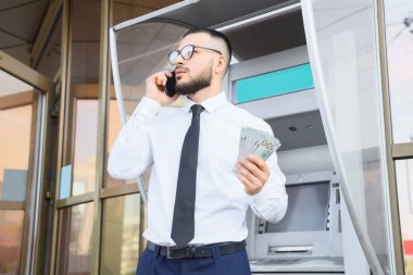 Dolar banknotları olan bir işadamı ATM 'nin yanında cep telefonuyla konuşuyor.